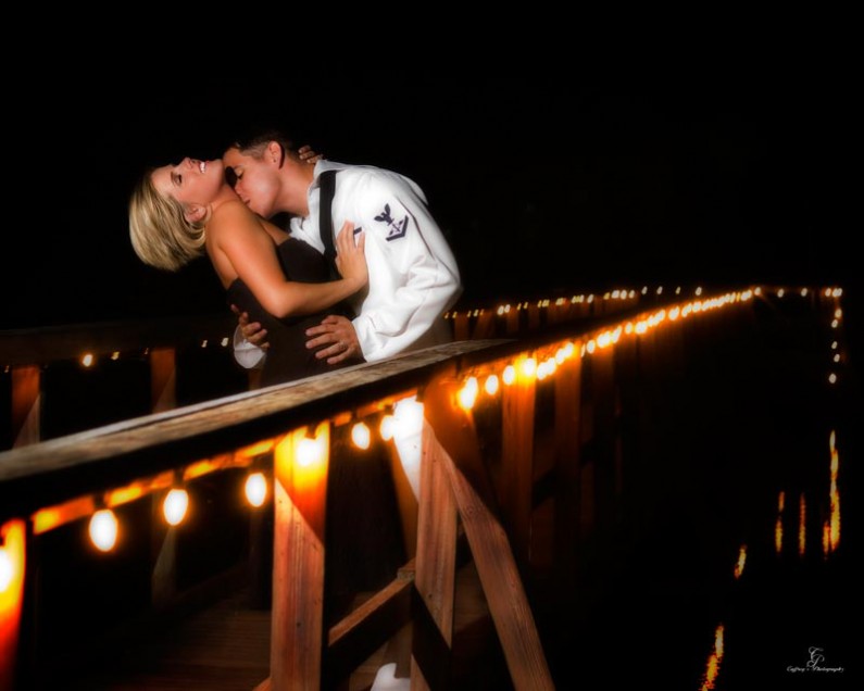 Sailor and his girl kissing on the lakeside deck thta is lit up with lights at night. 