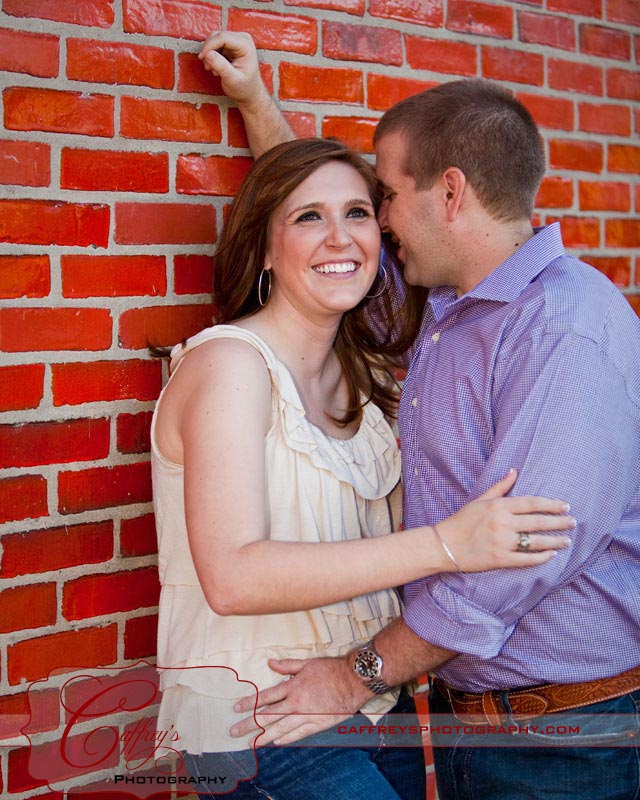 Groom whispering into his fiances ear next to a red wall during their engagment session. 